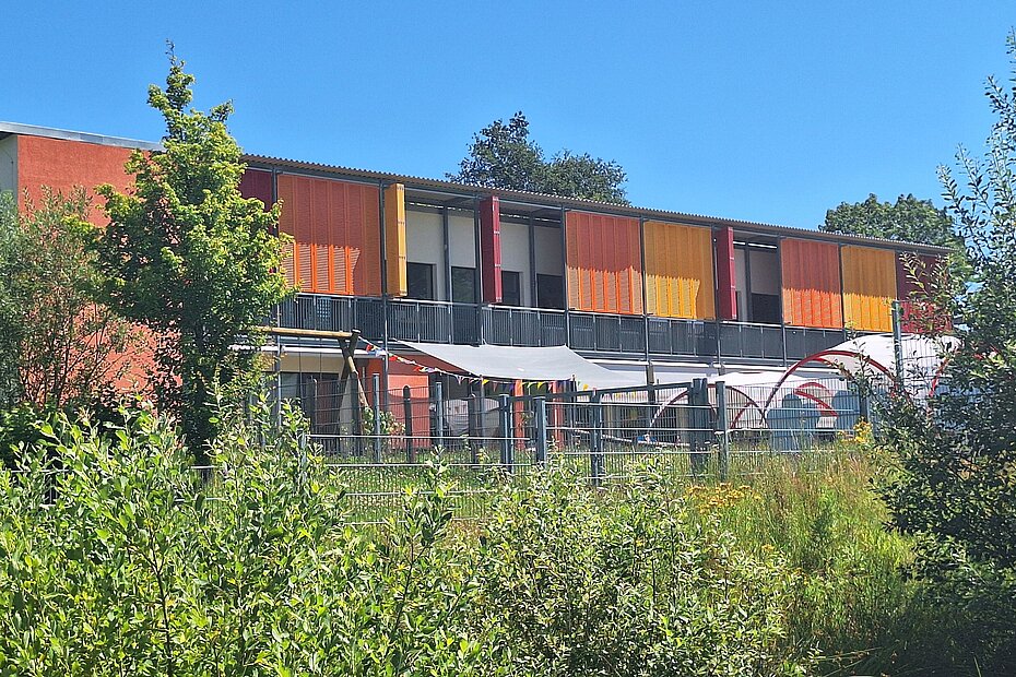 Das Foto zeigt eine klimaangepasste Kindertagesstätte in Rheinland-Pfalz. Es handelt sich um einen Pavillon mit Verschattung mit Holzläden. Im Vordergrund eine Hecke.