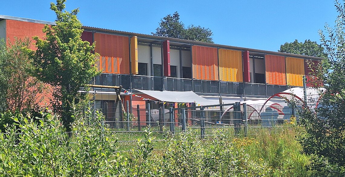 Das Foto zeigt eine klimaangepasste Kindertagesstätte in Rheinland-Pfalz. Es handelt sich um einen Pavillon mit Verschattung mit Holzläden. Im Vordergrund eine Hecke.