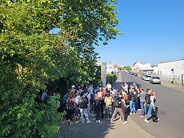 Eine Exkursionsgruppe steht im Gewerbegebiet zwischen Straße und Bäumen